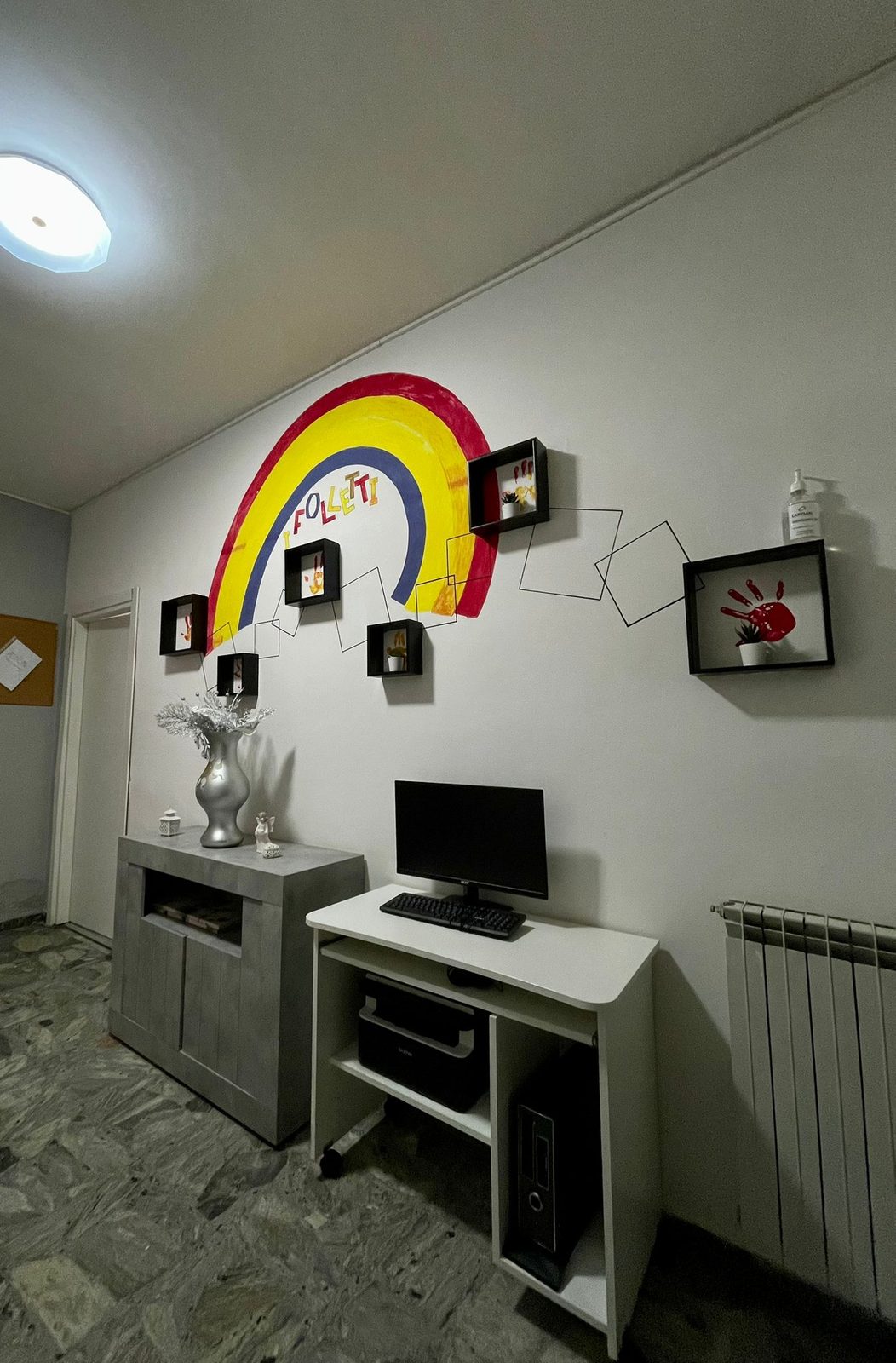 Corridoio con murale arcobaleno, cornici con impronte colorate e scrivania nella comunità I Folletti